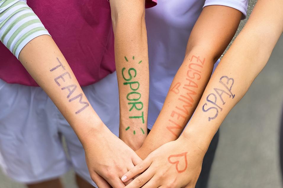Vier Arme liegen übereinander, mit farbiger Schrift: „TEAM“, „SPORT“, „ZUSAMMEN“, „SPASS“ auf der Haut.