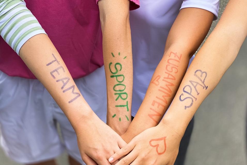 Vier Arme liegen übereinander, mit farbiger Schrift: „TEAM“, „SPORT“, „ZUSAMMEN“, „SPASS“ auf der Haut.